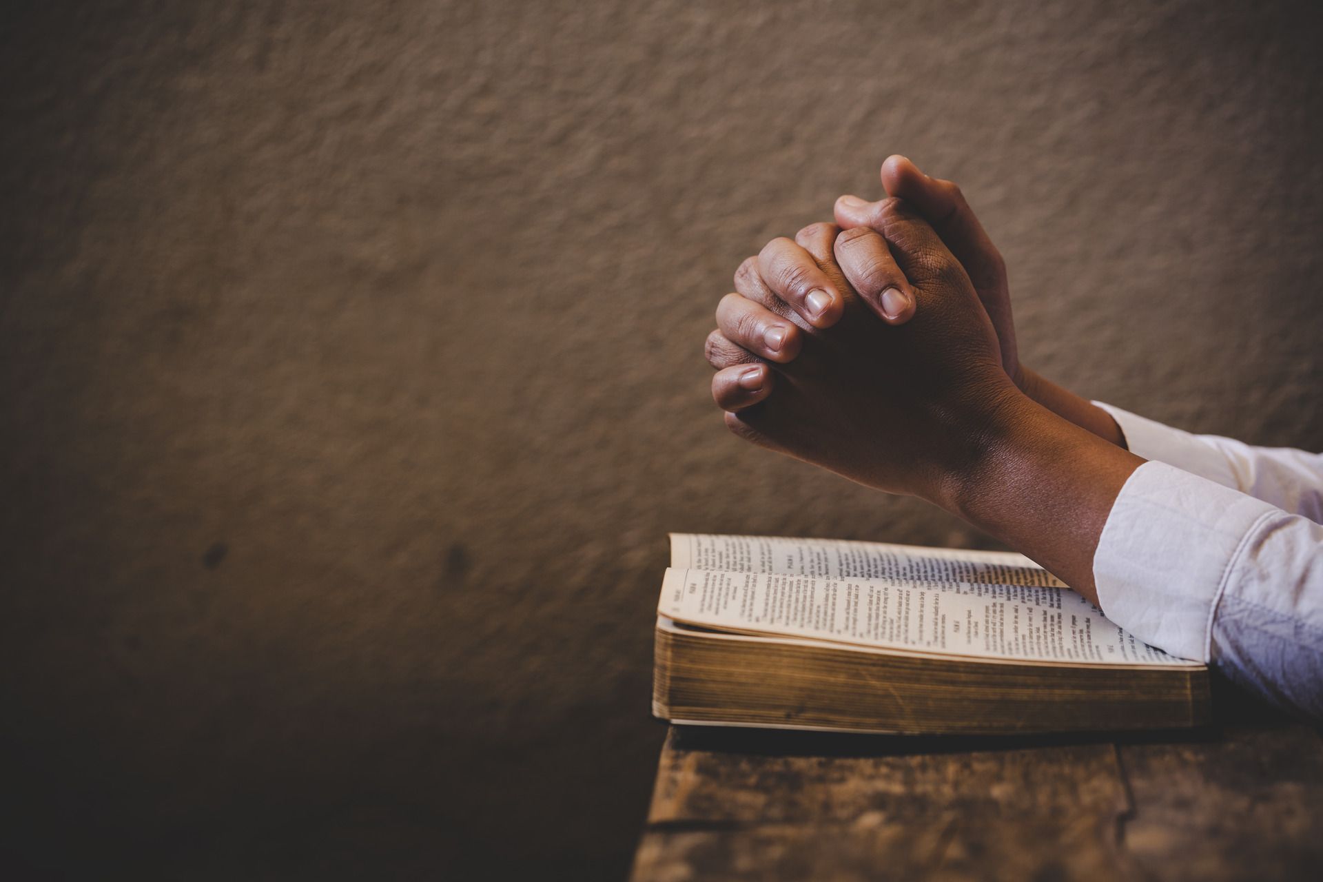 Hands clasped in prayer over an open book, resting on a wooden surface.