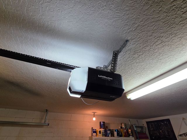 Black garage door opener mounted on a textured ceiling beside a fluorescent light and wall shelves.