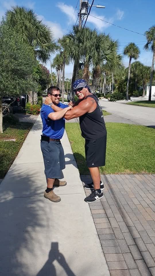 Two men greeting with a shoulder clasp on a sunny sidewalk near palm trees.