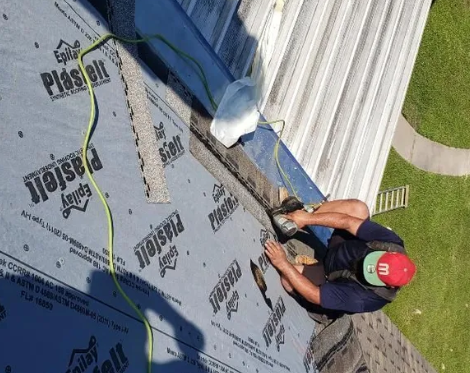 Worker installing roofing underlayment on a sloped roof beside white metal panels.