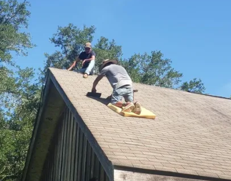 Two workers on a shingled roof, one kneeling and one standing, in bright daylight.