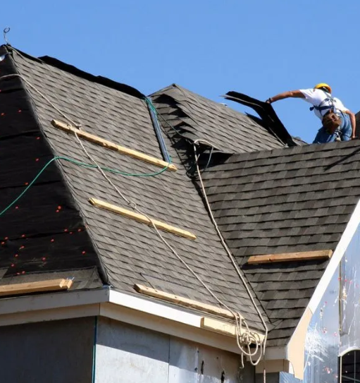 Worker repairing a dark shingled roof on a house under a clear blue sky