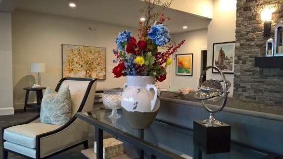 Lobby with a glass-topped table and floral arrangement. A chaise lounge is on the left; paintings and stone wall are in background.