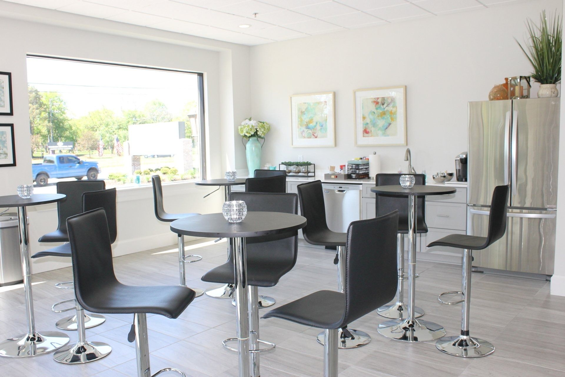 Lounge area with round tables, black chairs, and a window overlooking a street. Includes a refrigerator and coffee station.