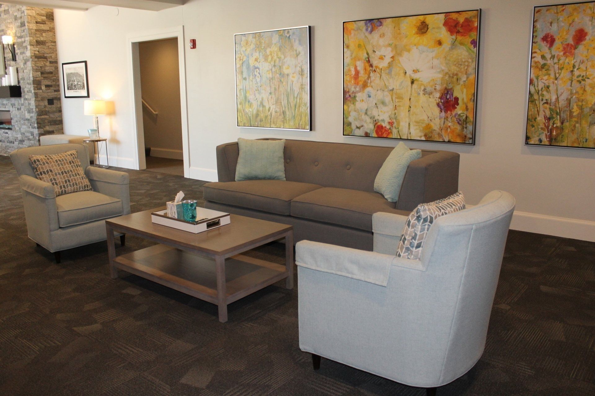 Cozy living room with gray couch, light blue armchairs, and colorful floral artwork. Coffee table with decorative items.