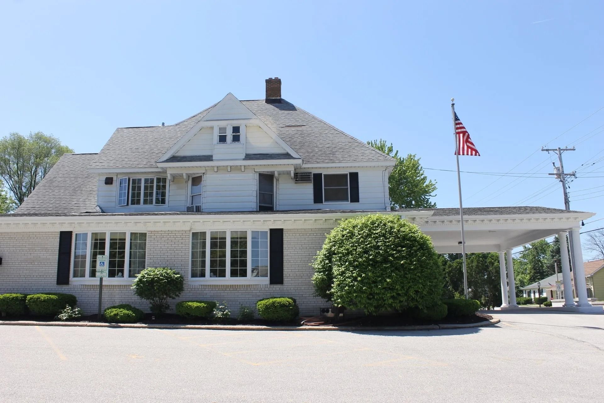 White building with a brick facade and black shutters, American flag flying, shrubs in front, bright sunny day.