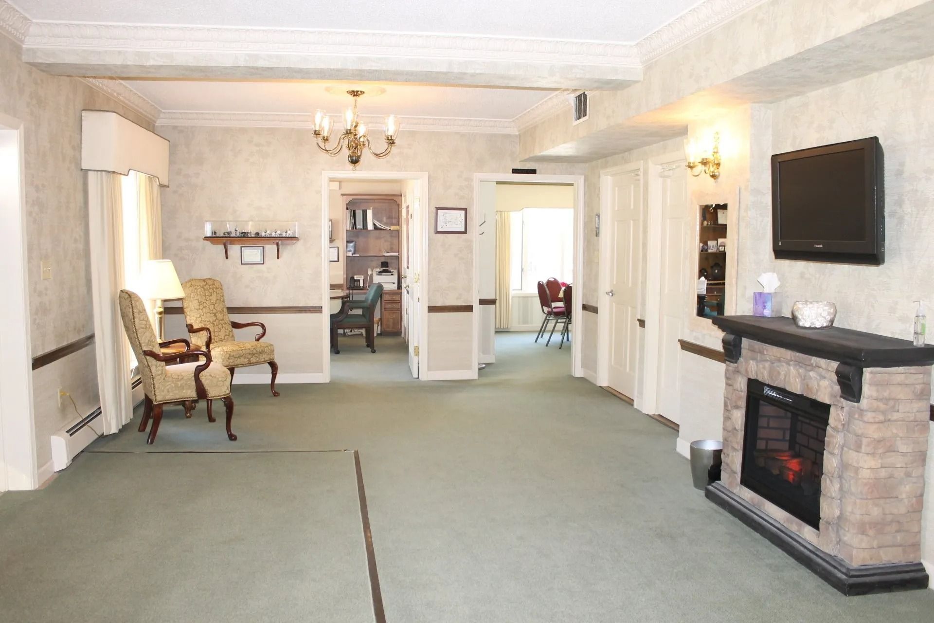 Waiting room with fireplace, chairs, and doorways. Green carpet, neutral walls, and a mounted TV.