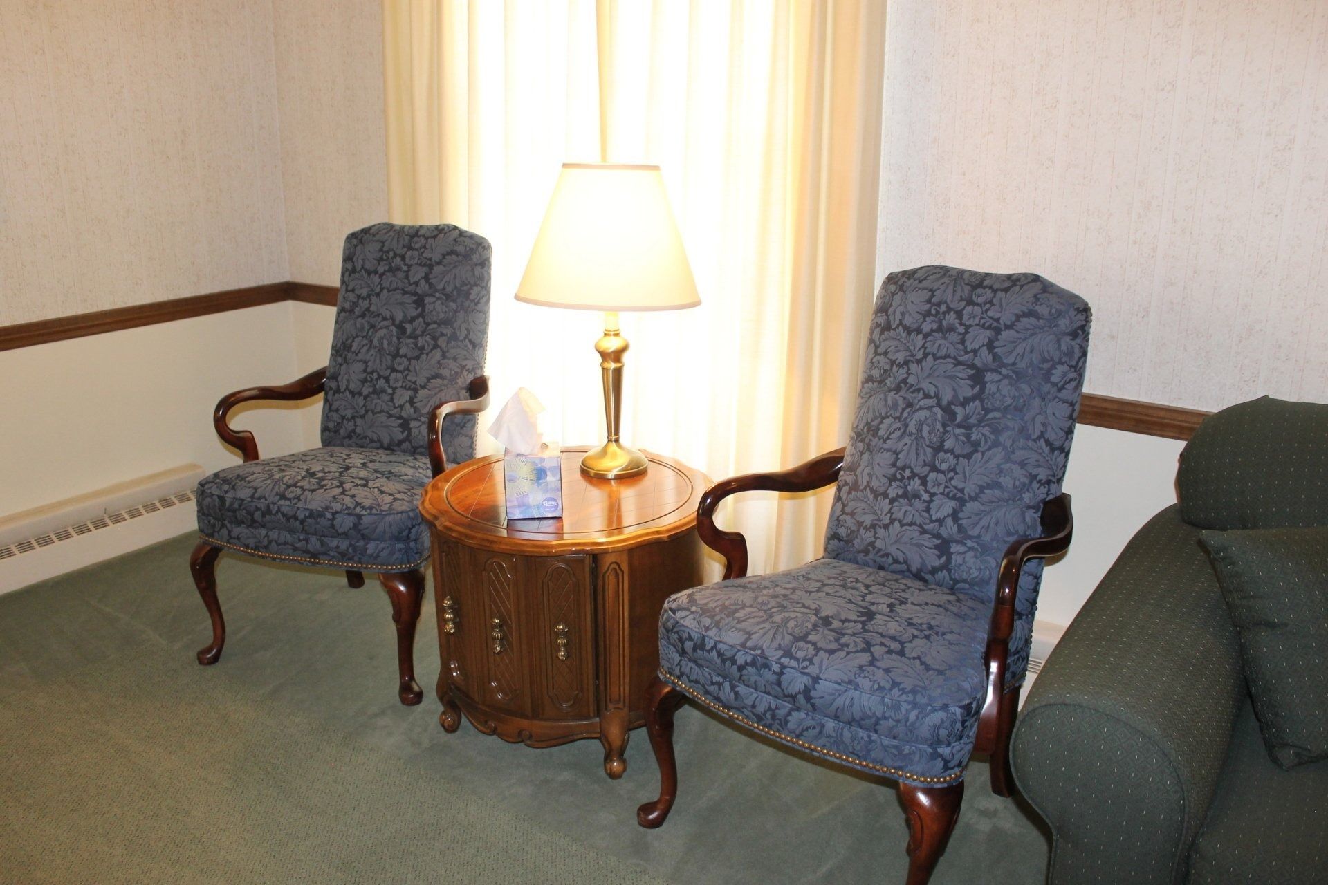 Two upholstered blue chairs and a lamp-lit table in a room with green carpet and a window.