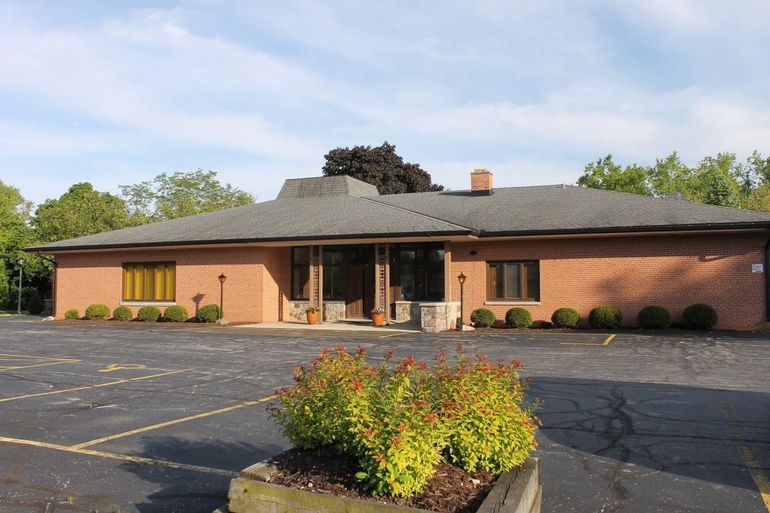 Brick building with dark roof, parking lot, and landscaping on a sunny day.