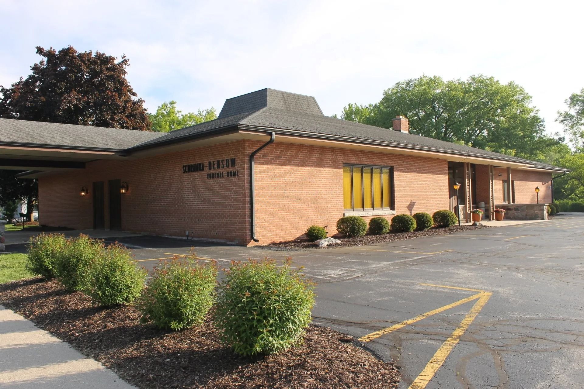 Brick building with dark roof, bushes, and a sign that reads