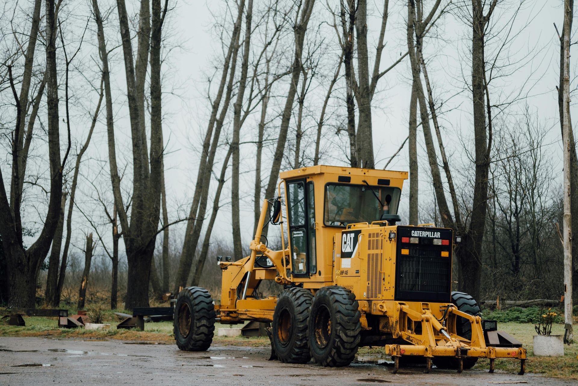 Yellow road grader parked on a damp road amid bare trees in a cloudy setting.