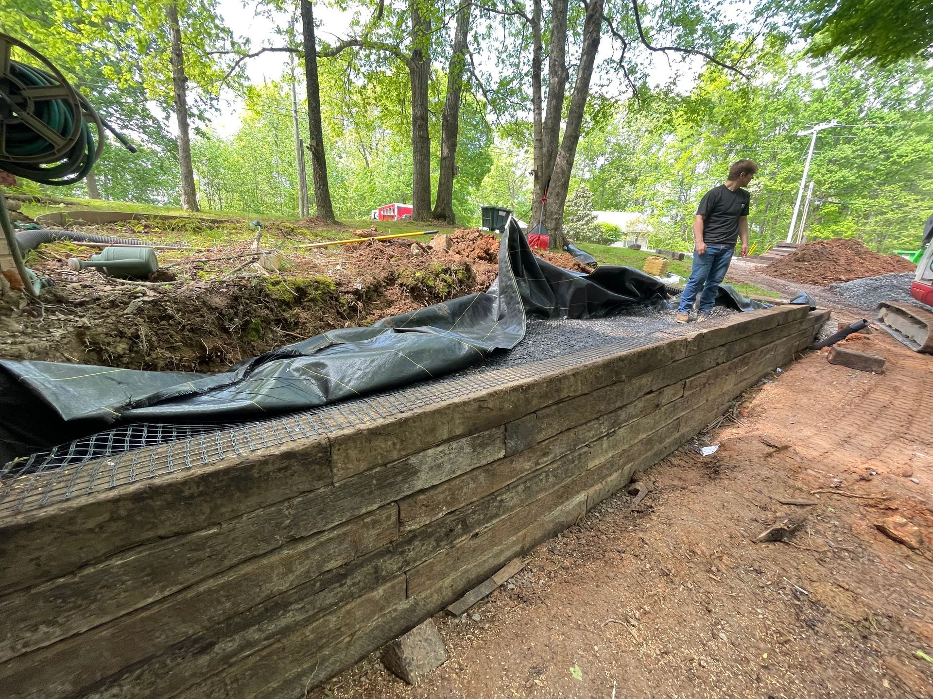 A man standing near a retaining wall made of stacked wood. Dirt and trees are in the background.