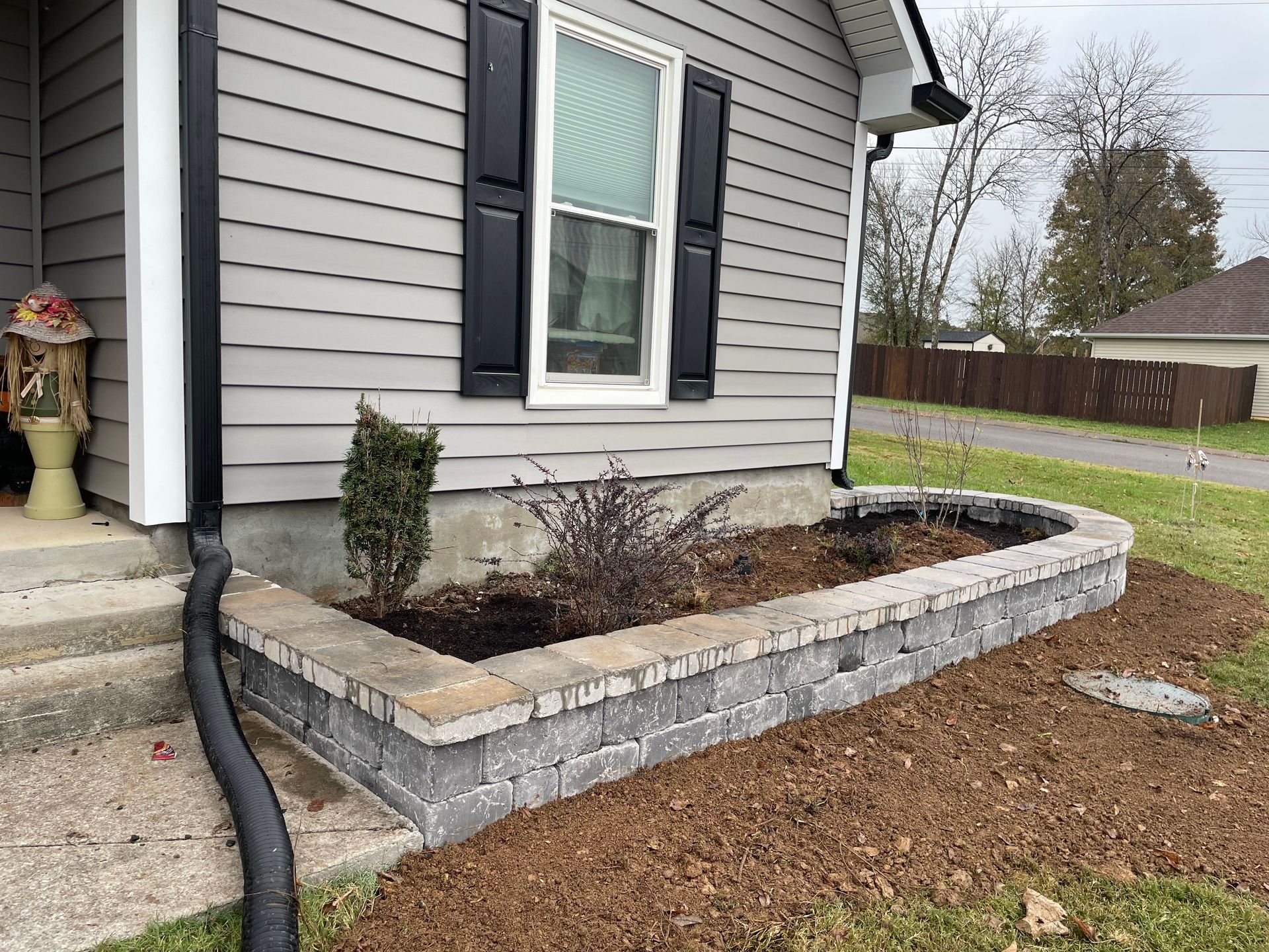 Gray house with stone flower bed, black shutters, and downspout on a grassy lawn.