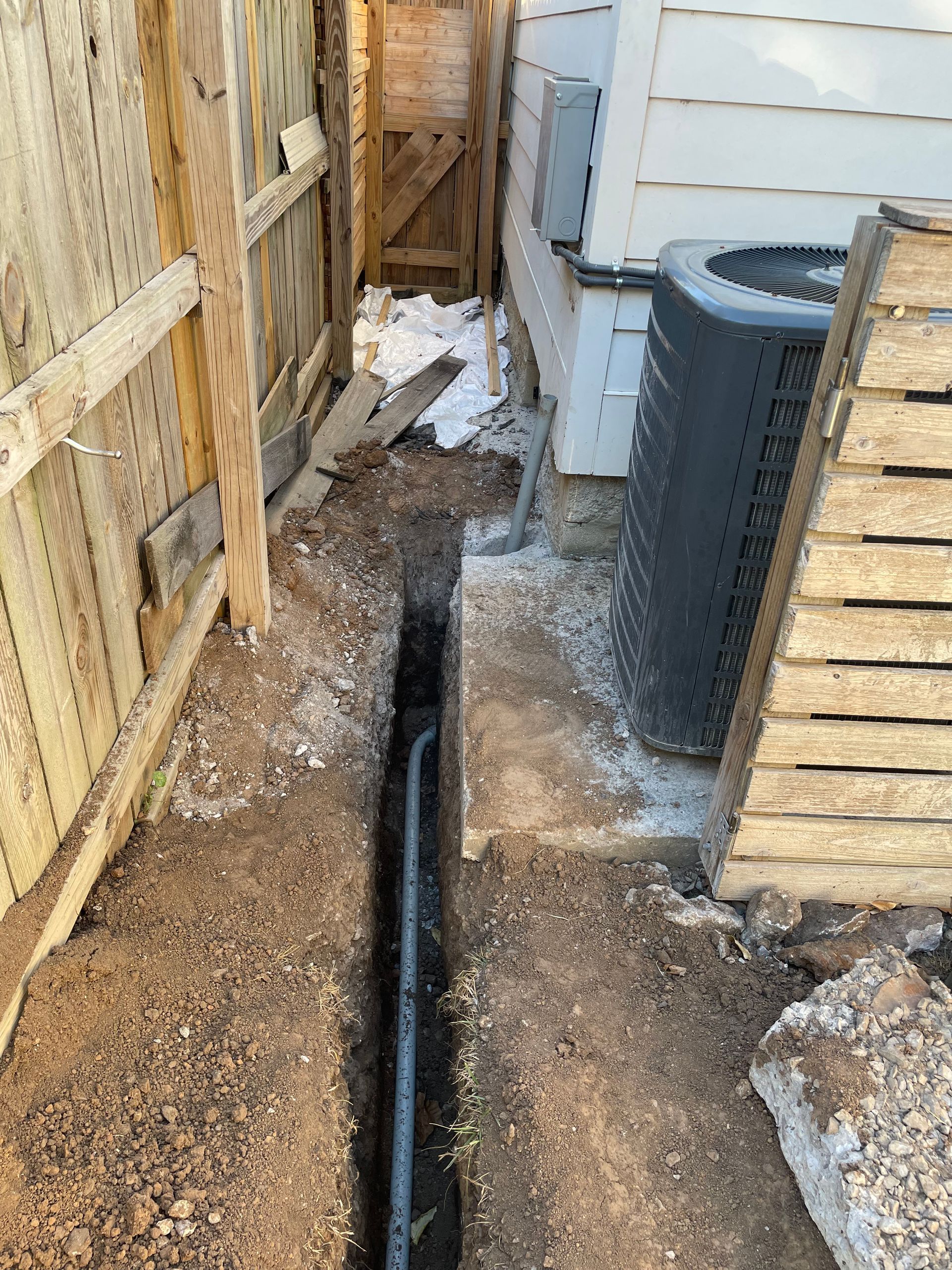 A hole in the ground next to a house with an air conditioner.
