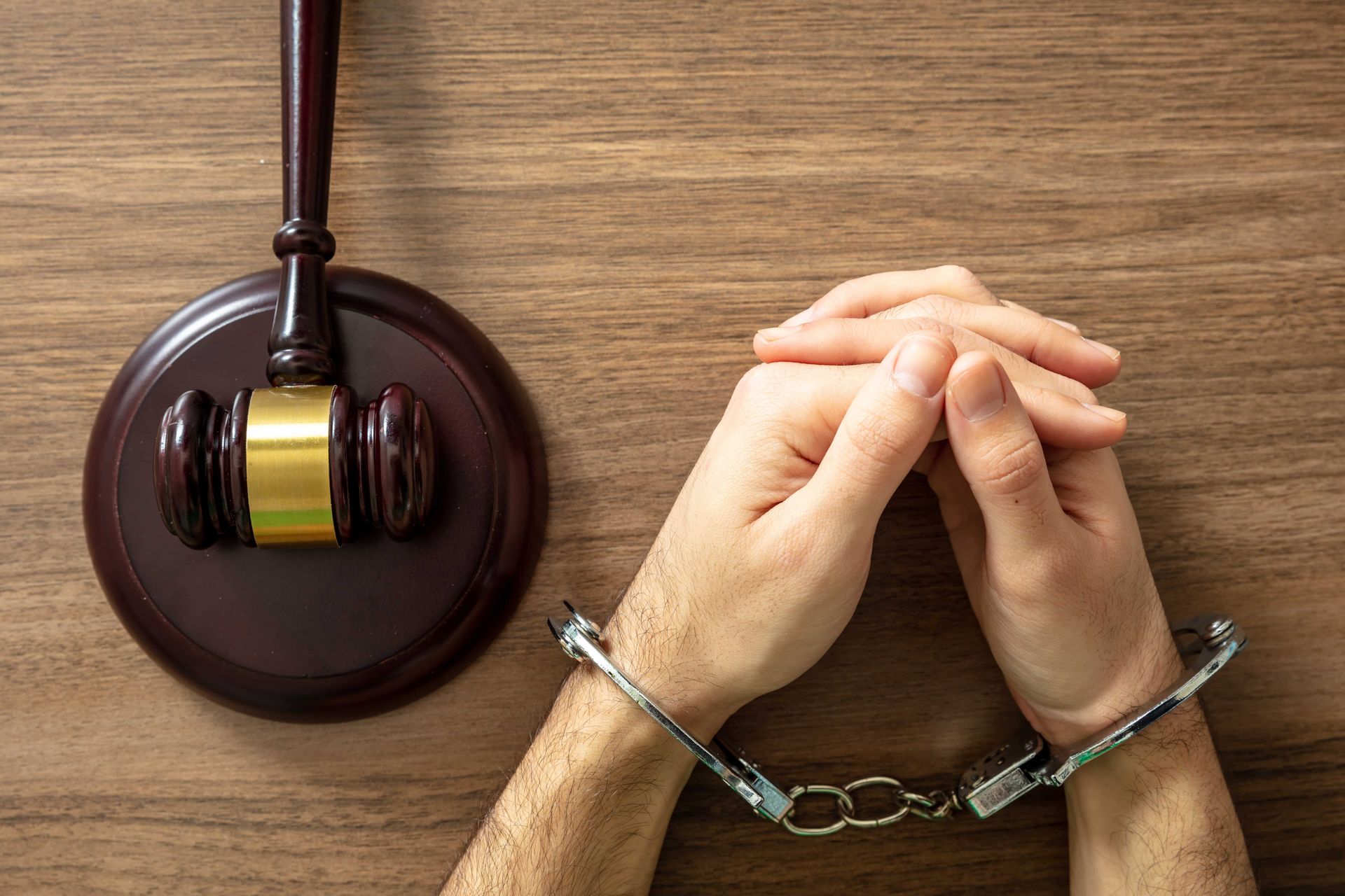 Handcuffed hands next to a gavel on a wooden surface.