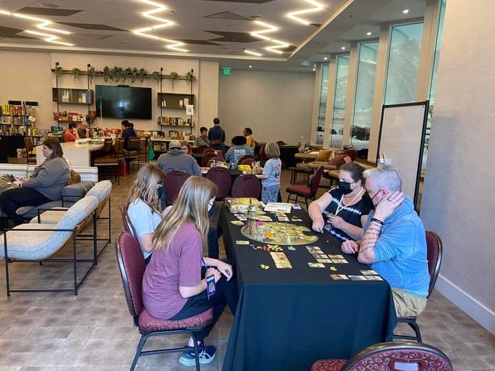 People playing board games at tables in a brightly lit room. Other people are in the background.
