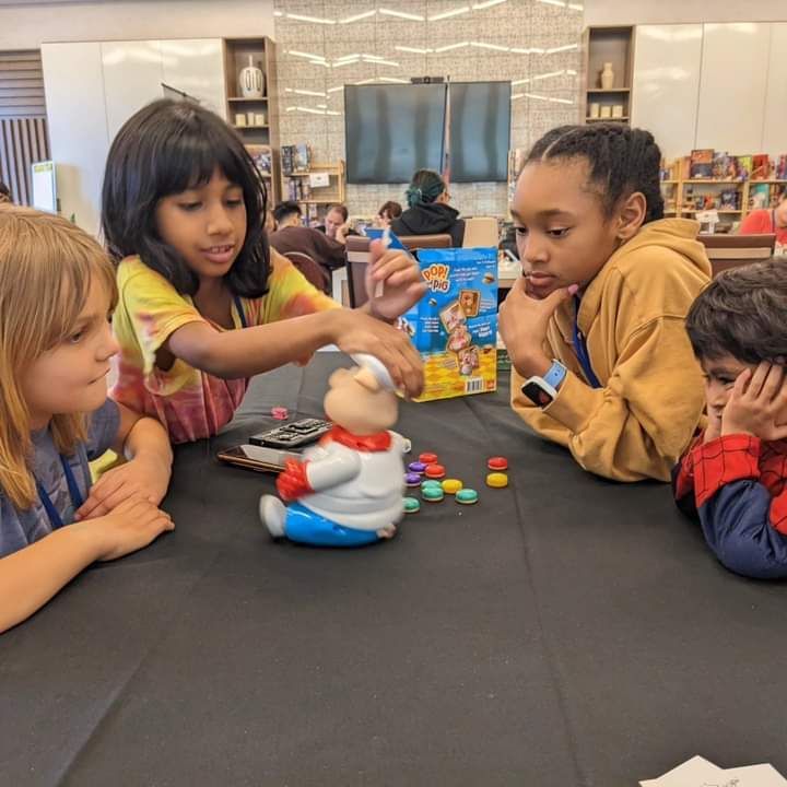 Children playing a board game at a table. One child is placing a token. Others watch intently.