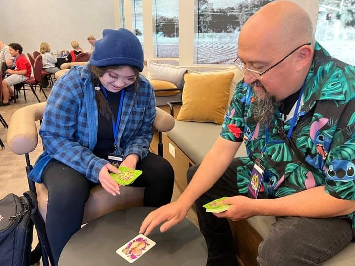 Man and teen with a blue hat play cards at a table indoors. The person smiles.