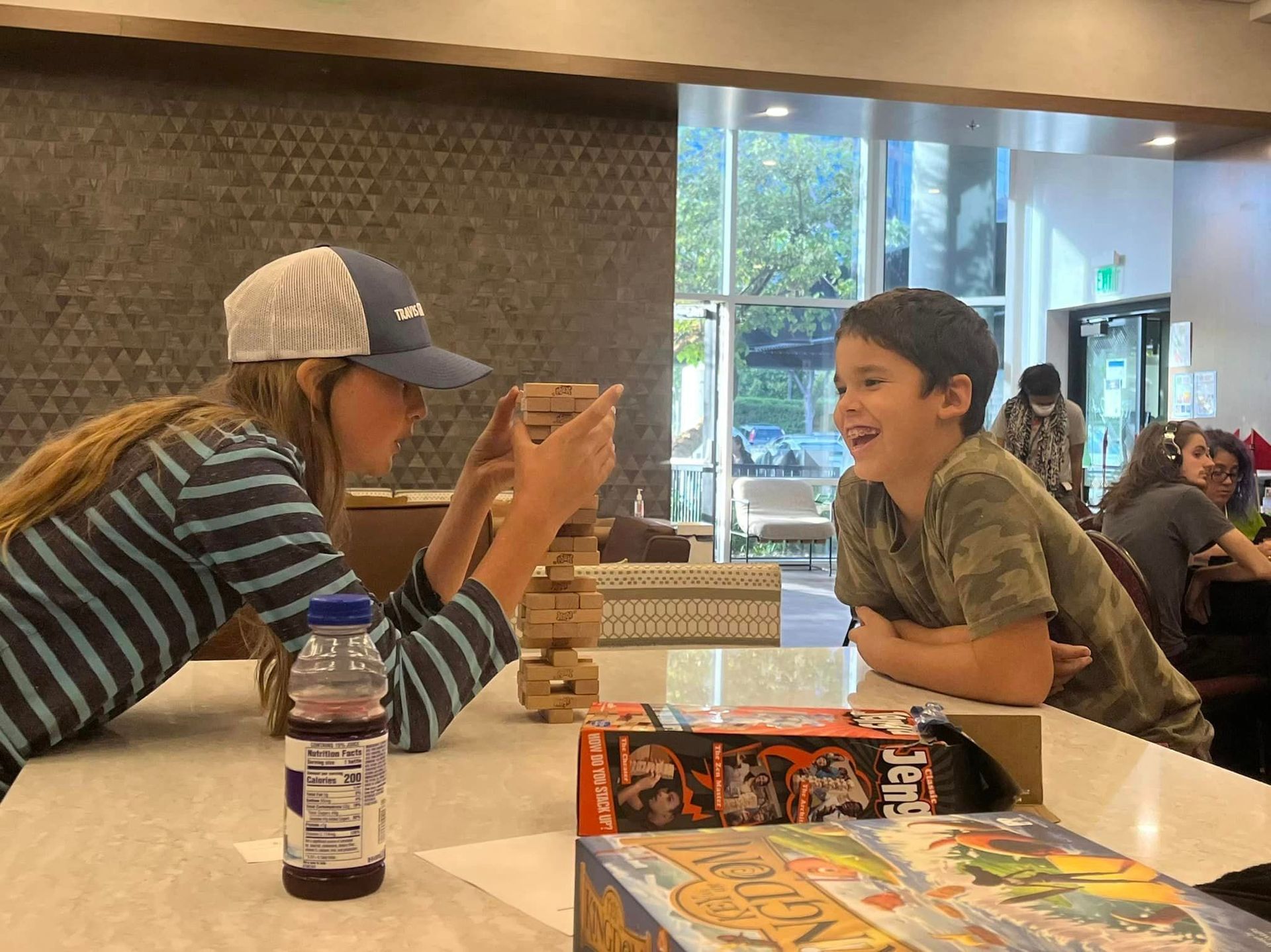 Two people playing Jenga, one smiling, in a brightly lit room. A drink and board game are on the table.