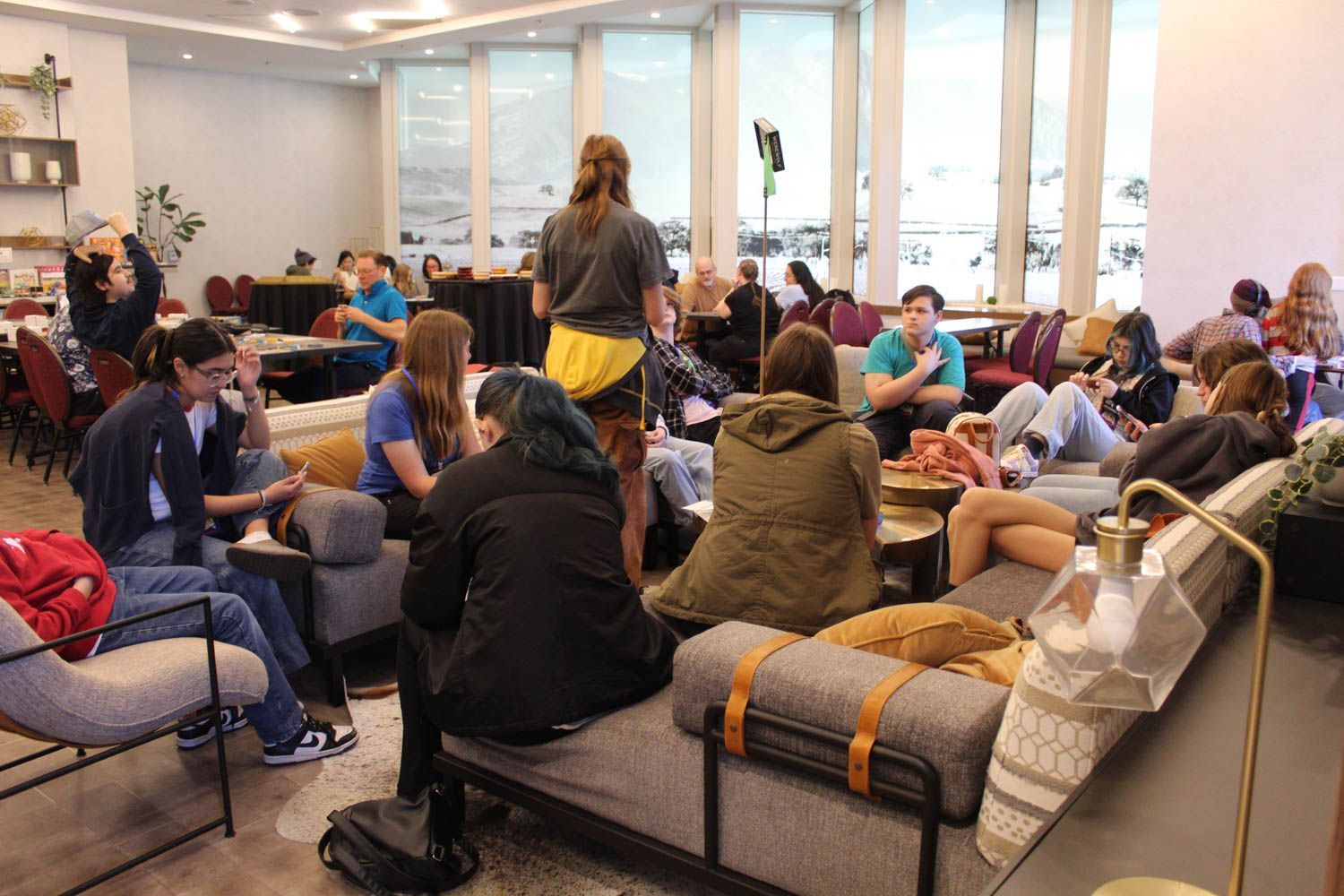 People gathered in a well-lit lobby with large windows; some seated on couches, others standing, engaging in conversation.