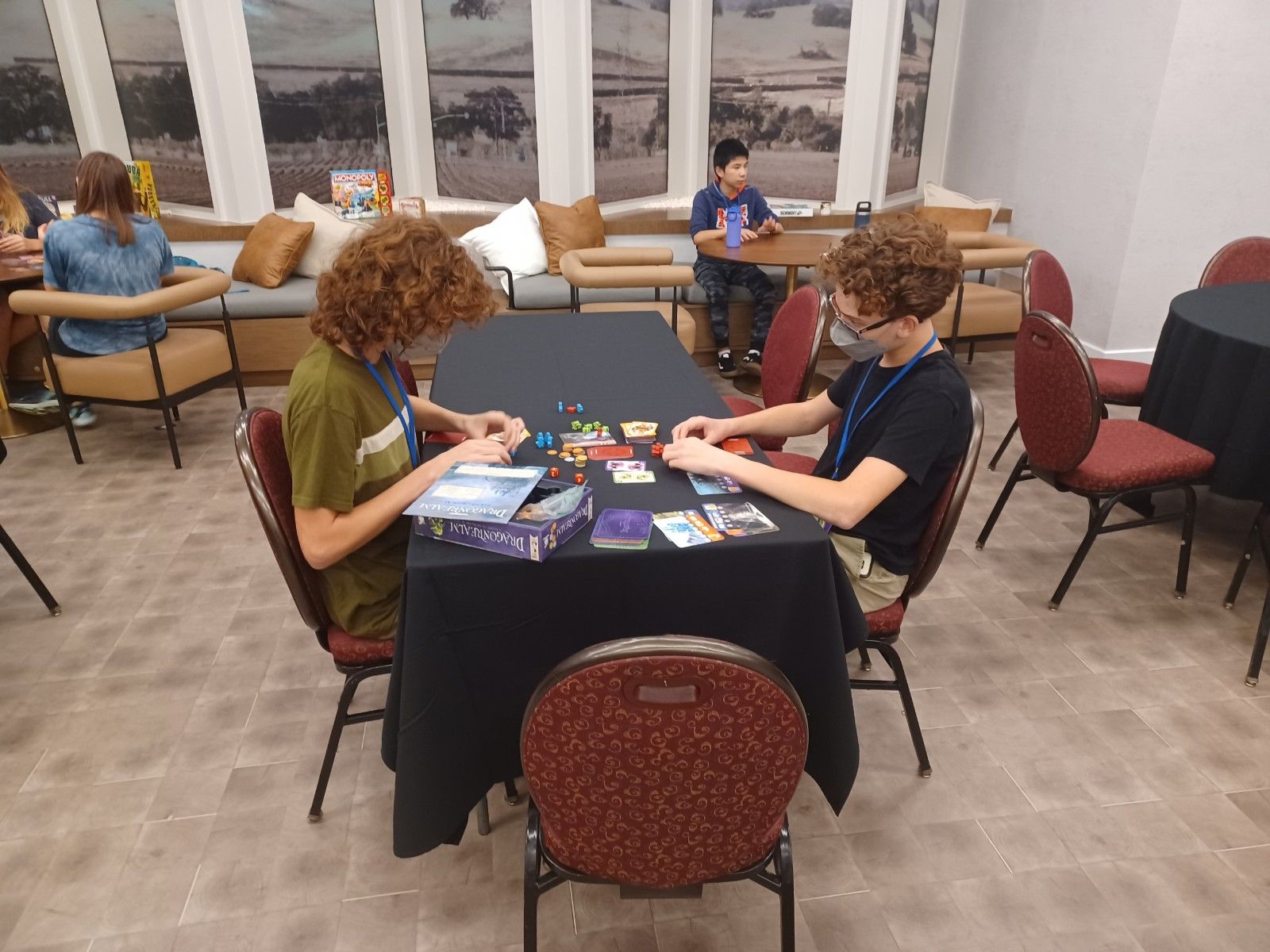 Two people sit at a table playing a board game; others are in a room with a window, couches, and tables.