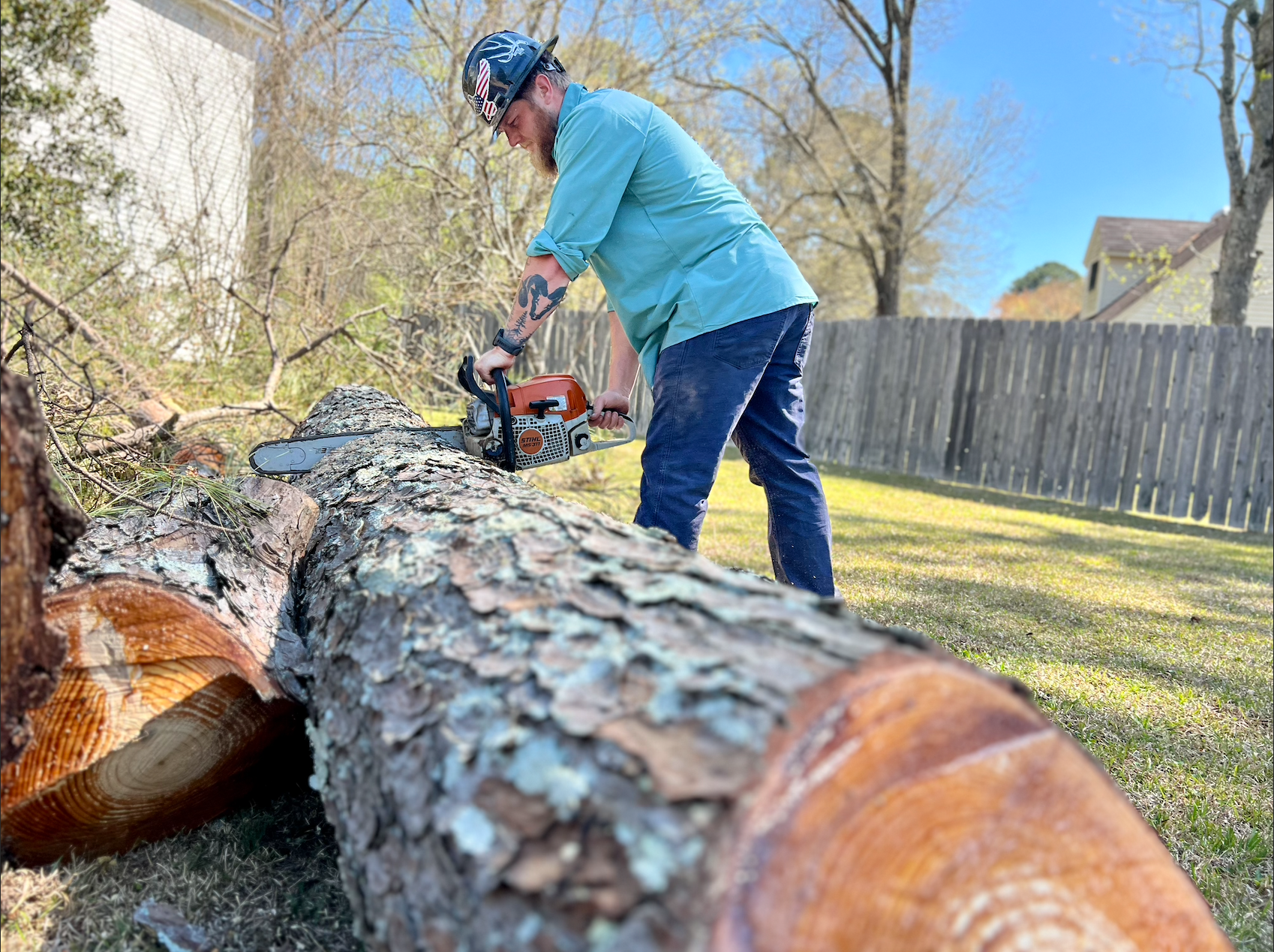 Man using a chainsaw to cut a log in a grassy backyard, wearing safety gear, blue shirt, and jeans.