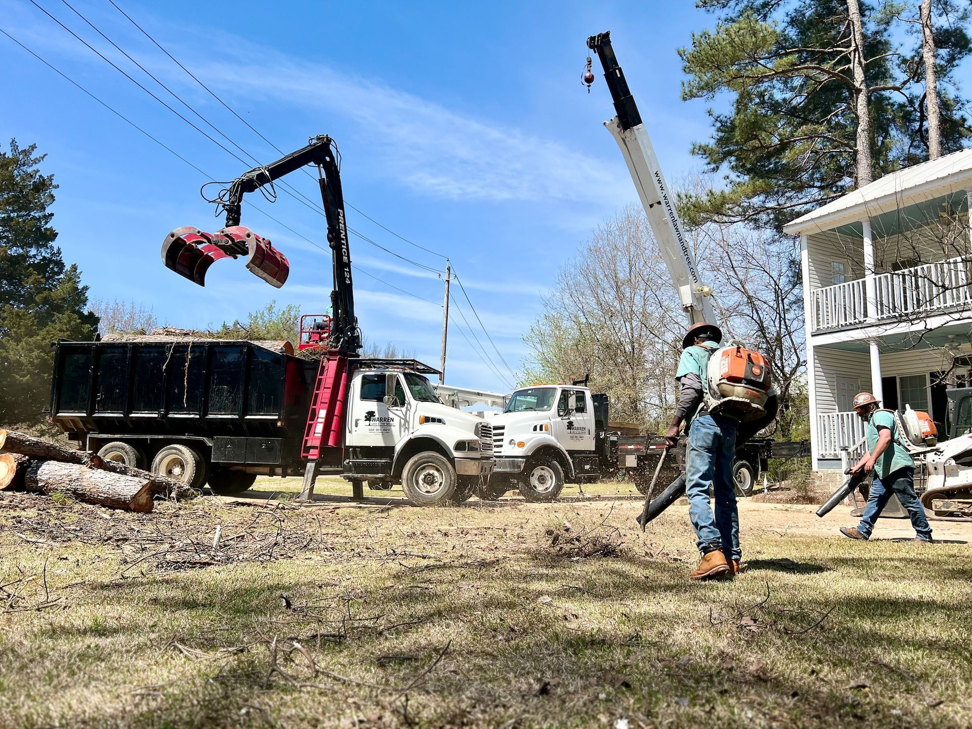 Tree removal: Workers using equipment to load debris into a dump truck, with a house in the background.