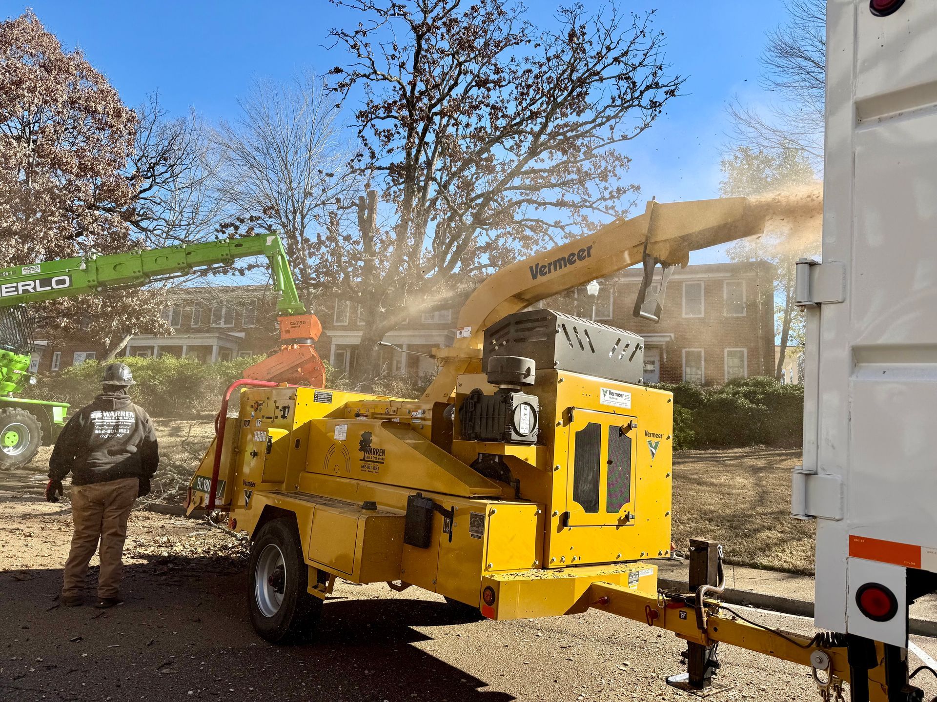 A yellow wood chipper shreds branches; a worker in safety gear stands nearby; green lift truck feeds it.