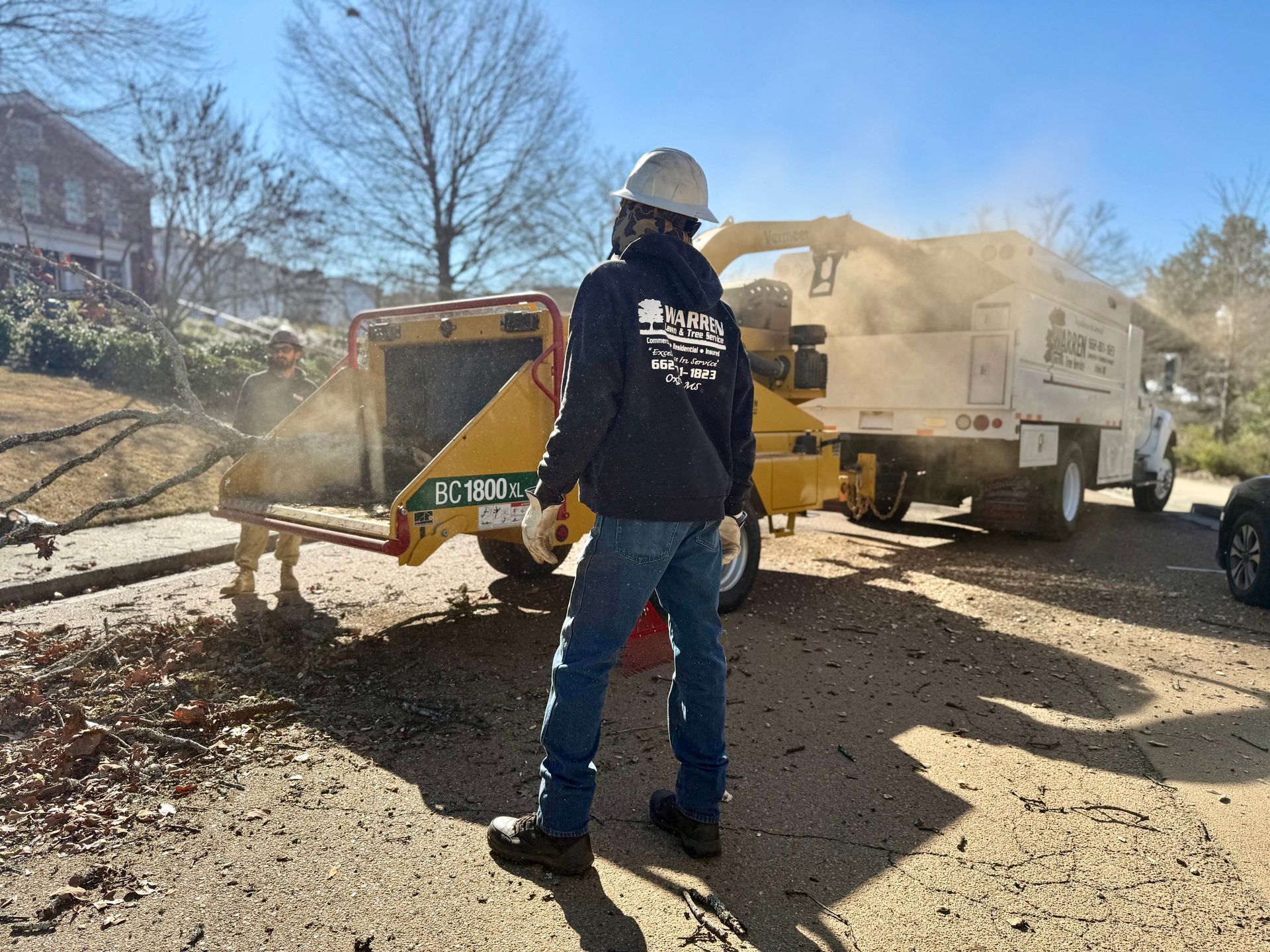 Arborist wearing helmet, feeding branches into a wood chipper; truck in background.