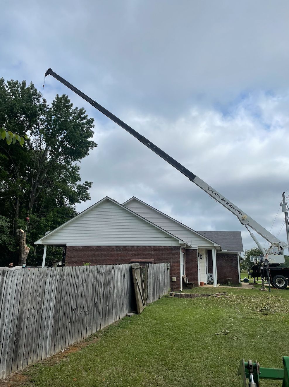 A tall crane reaching over a brick house with a wooden fence in the foreground. Overcast sky.