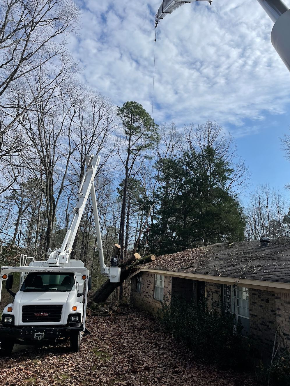White utility truck with an extended arm, trimming a tree that has fallen on a house. Overcast sky.