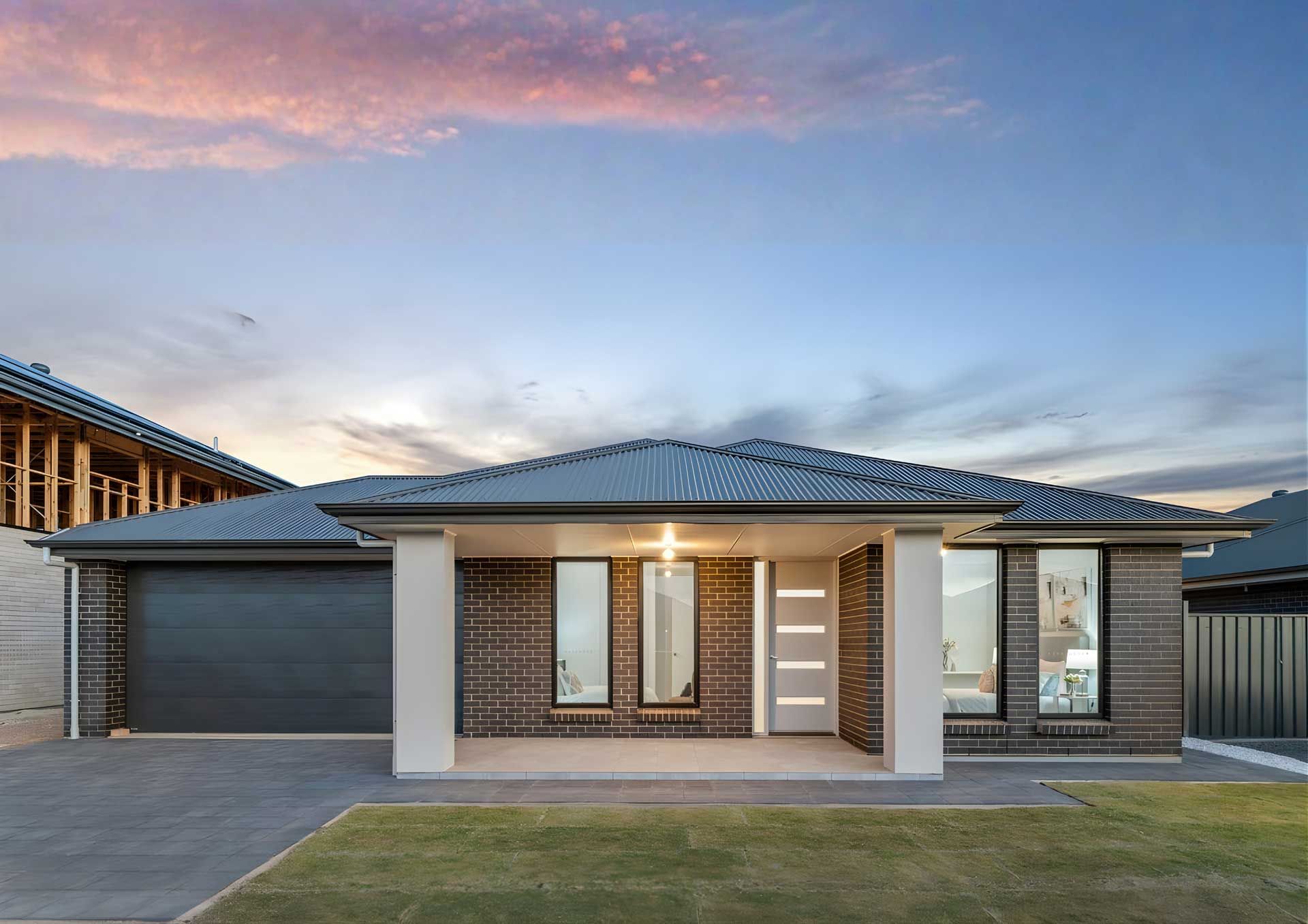 Modern brick home with gray garage door, columns, and windows, set against a sunset sky.