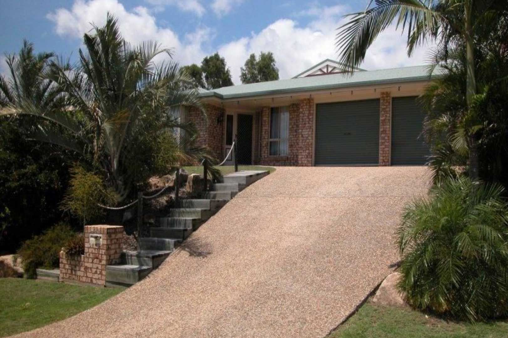 House with brown brick exterior, tan driveway leading to three-car garage, steps to the front door, blue sky, palm trees.