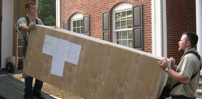 Two men are carrying a large piece of wood in front of a brick building from Cupboard Wagon cabinet delivery services