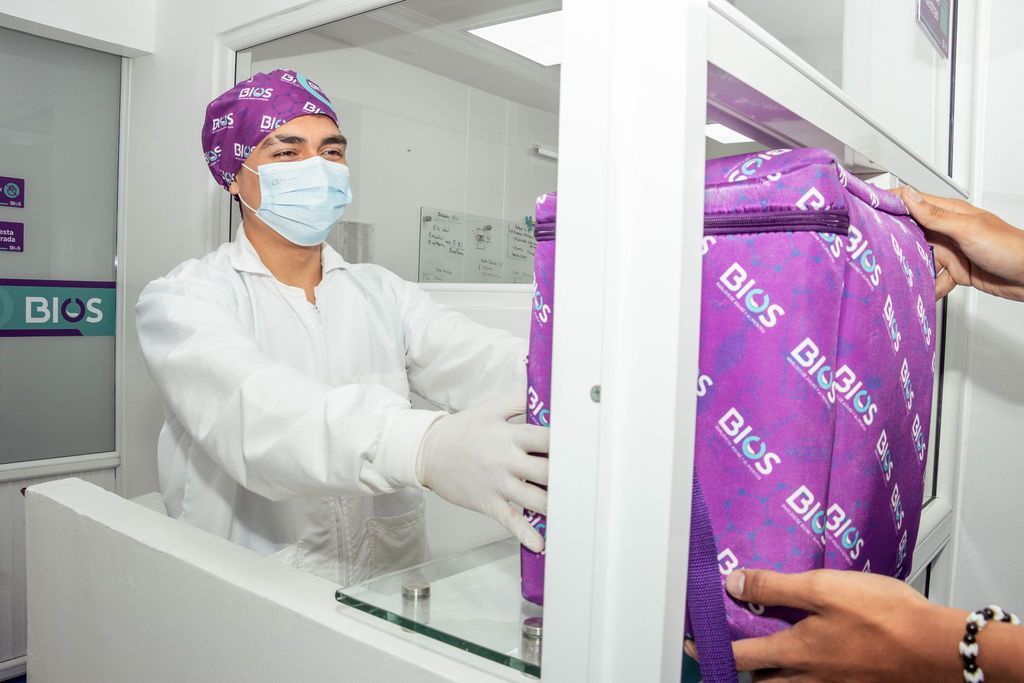 Un trabajador con bata blanca y mascarilla entrega un paquete morado a través de una ventanilla de atención al público.