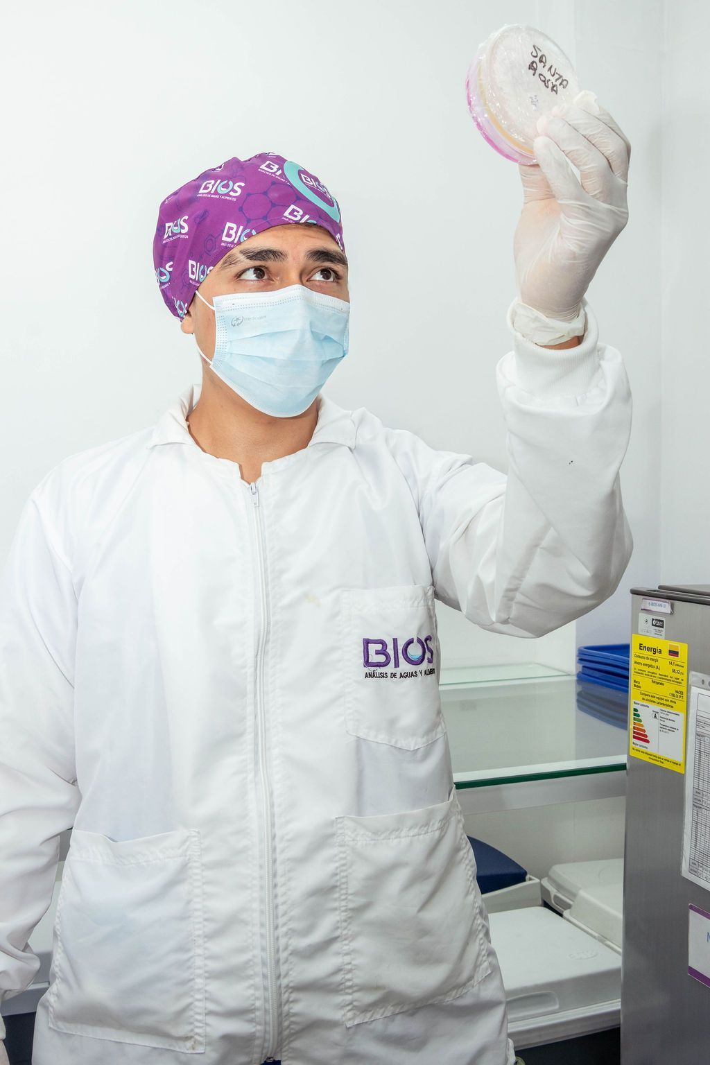 Un técnico de laboratorio con bata blanca y mascarilla sostiene una placa de Petri en un laboratorio clínico.