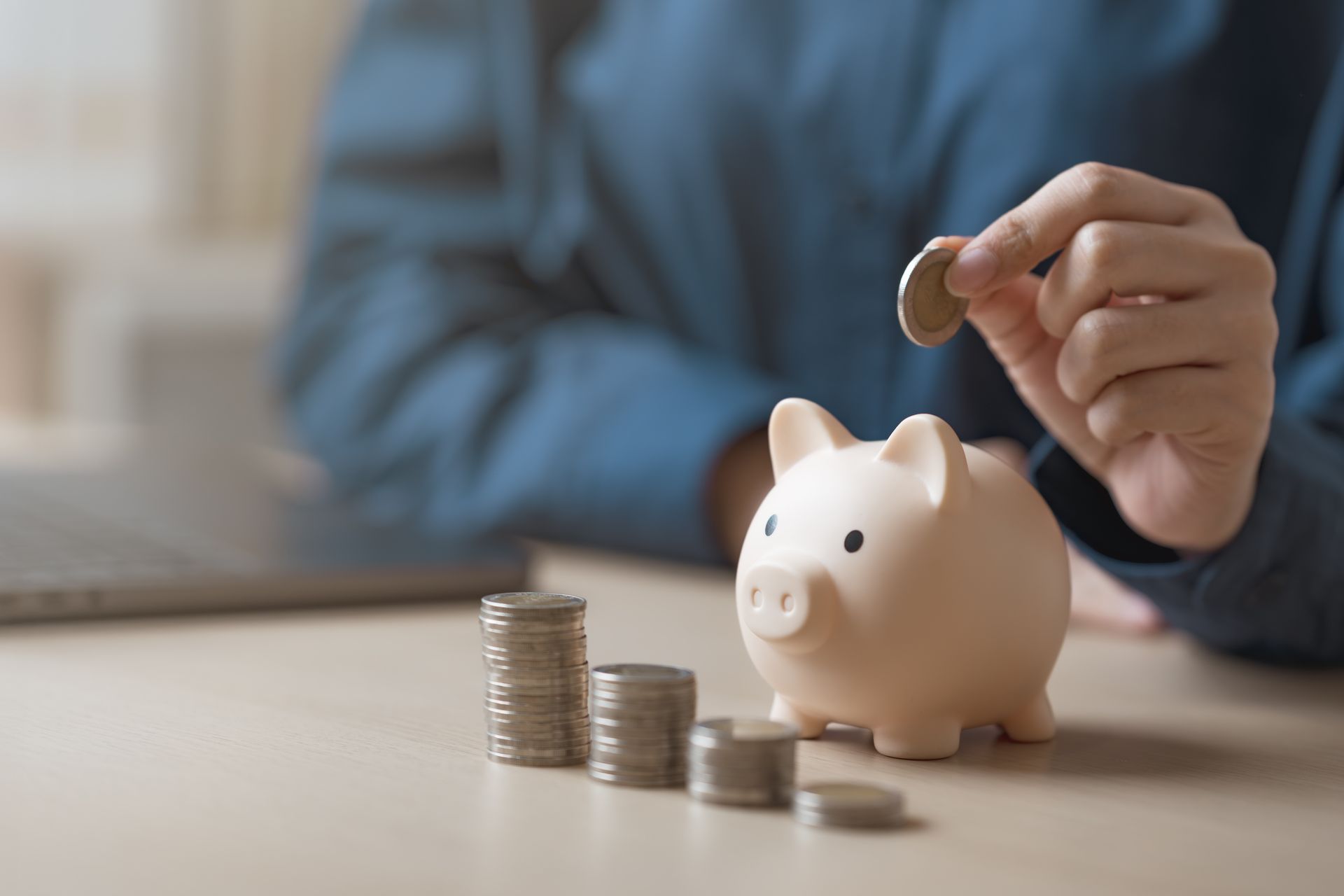 Person adding a coin to a piggy bank; stacks of coins on the table. Person adding a coin to a piggy bank; stacks of coins on the table.