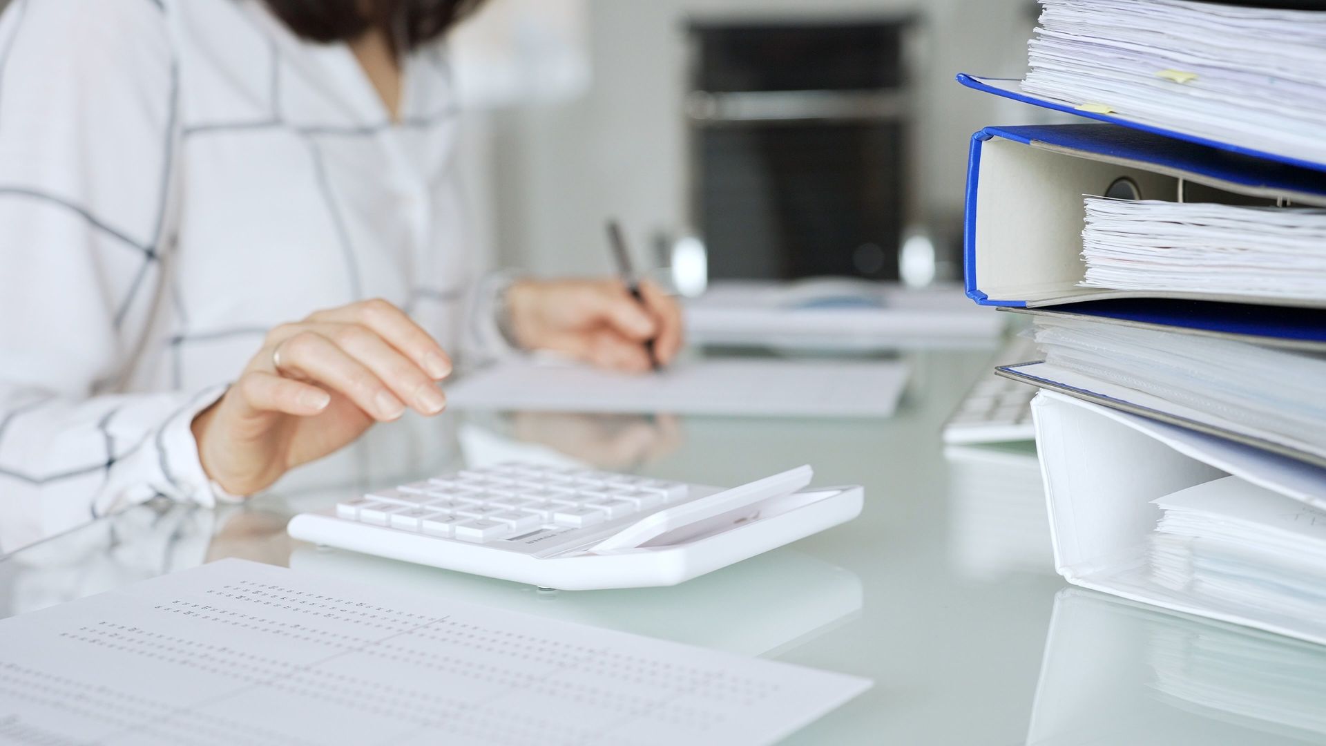 Person using a calculator, taking notes, with a stack of binders. Person using a calculator, taking notes, with a stack of binders.