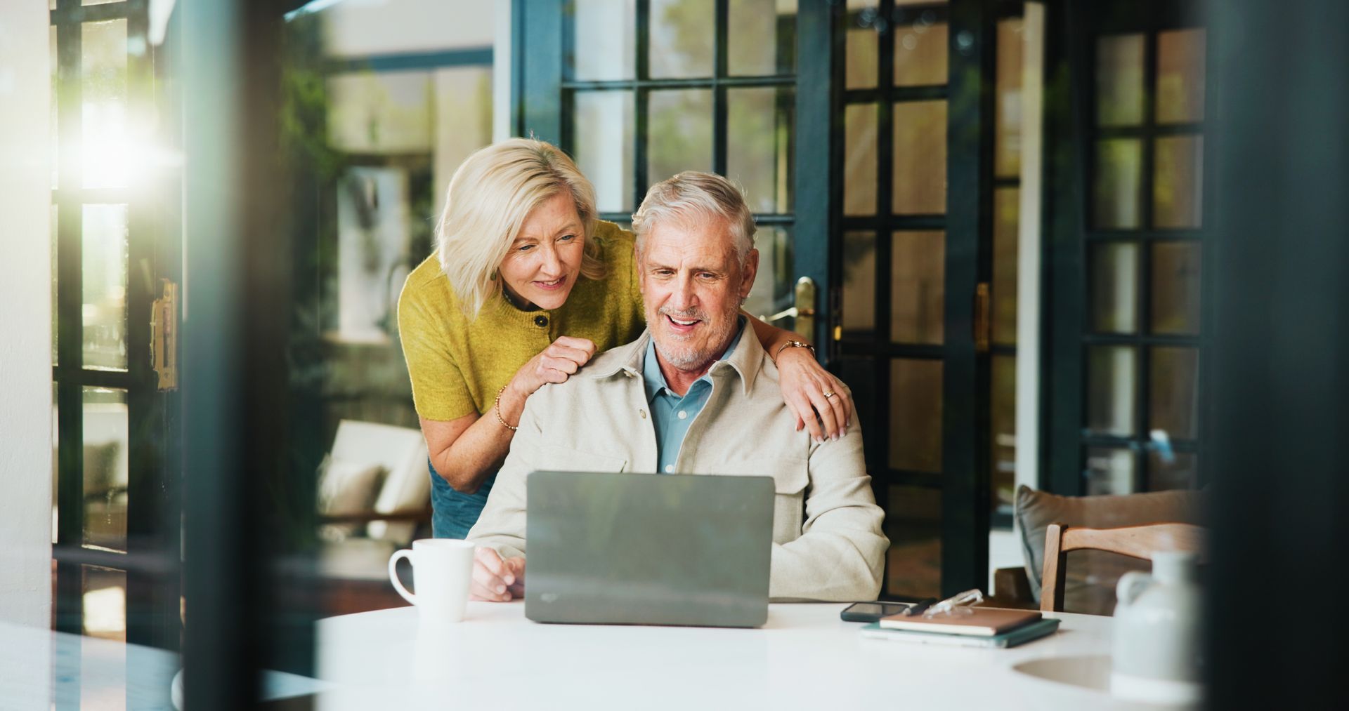 An older couple looks at a laptop together indoors. The woman has her arm around the man, smiling. An older couple looks at a laptop together indoors. The woman has her arm around the man, smiling.