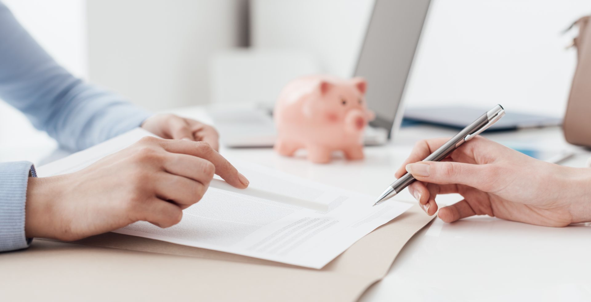Hands pointing to a document as another hand holds a pen, near a piggy bank and laptop. Hands pointing to a document as another hand holds a pen, near a piggy bank and laptop.