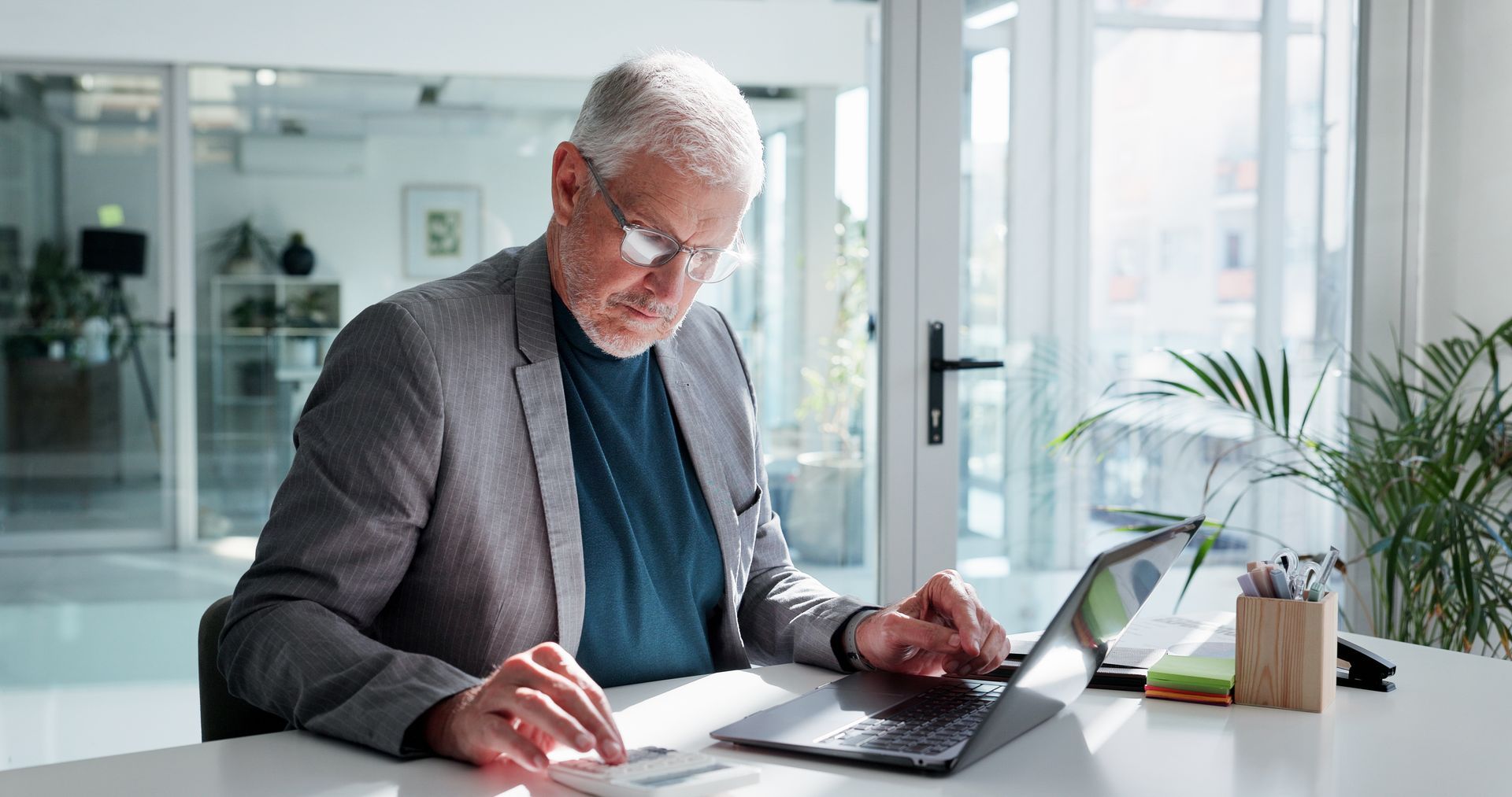 Man in a grey blazer works on a laptop at a white desk in a well-lit office. Man in a grey blazer works on a laptop at a white desk in a well-lit office.