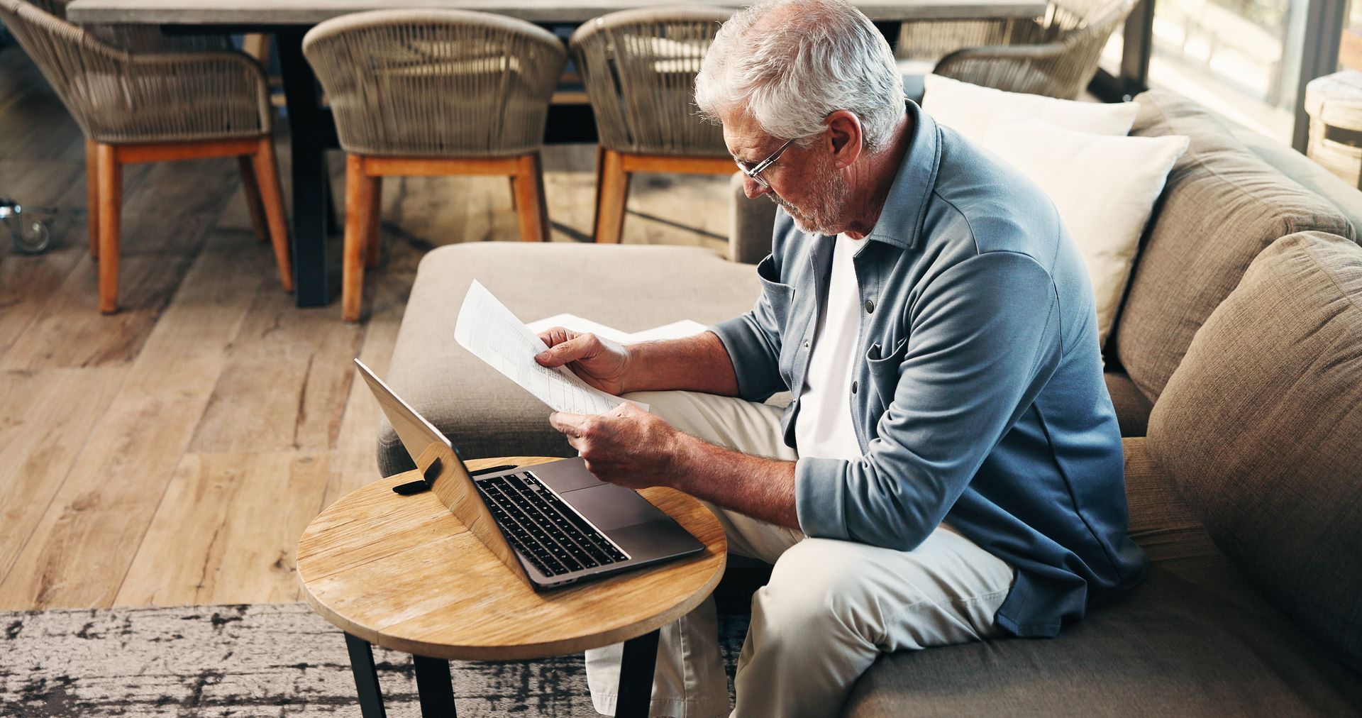 Senior man reviewing papers, using a laptop on a small table, sitting on a couch in a home setting. Senior man reviewing papers, using a laptop on a small table, sitting on a couch in a home setting.