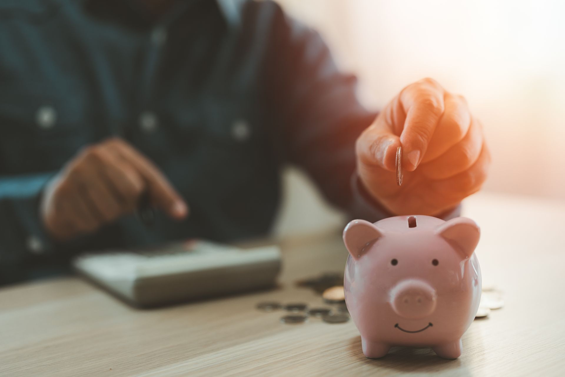 A hand dropping a coin into a pink piggy bank on a desk next to coins and a calculator. A hand dropping a coin into a pink piggy bank on a desk next to coins and a calculator.