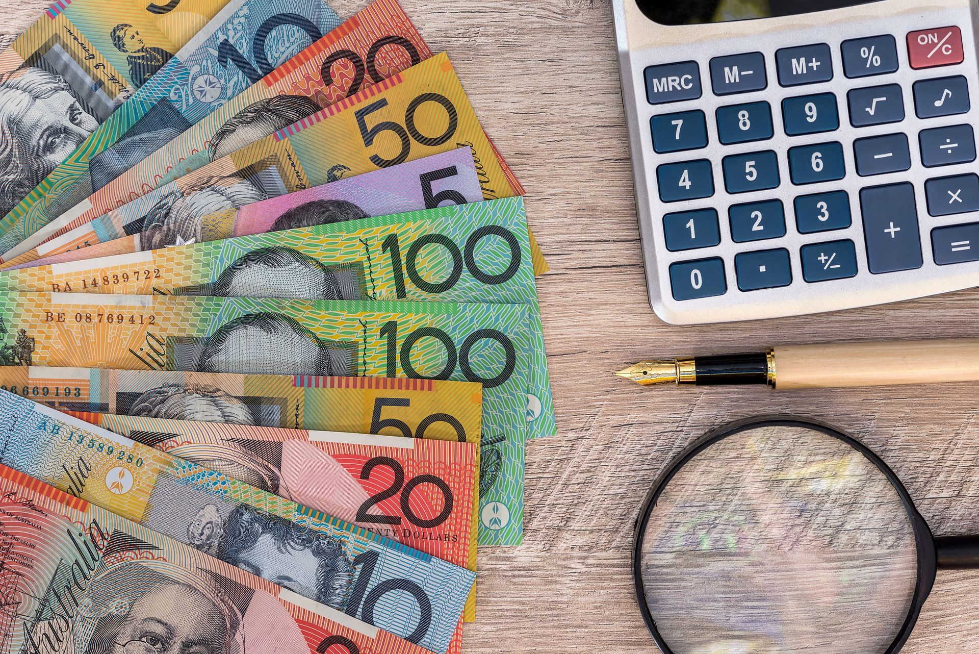Australian banknotes fanned out with a calculator, pen, and magnifying glass on wood. Australian banknotes fanned out with a calculator, pen, and magnifying glass on wood.