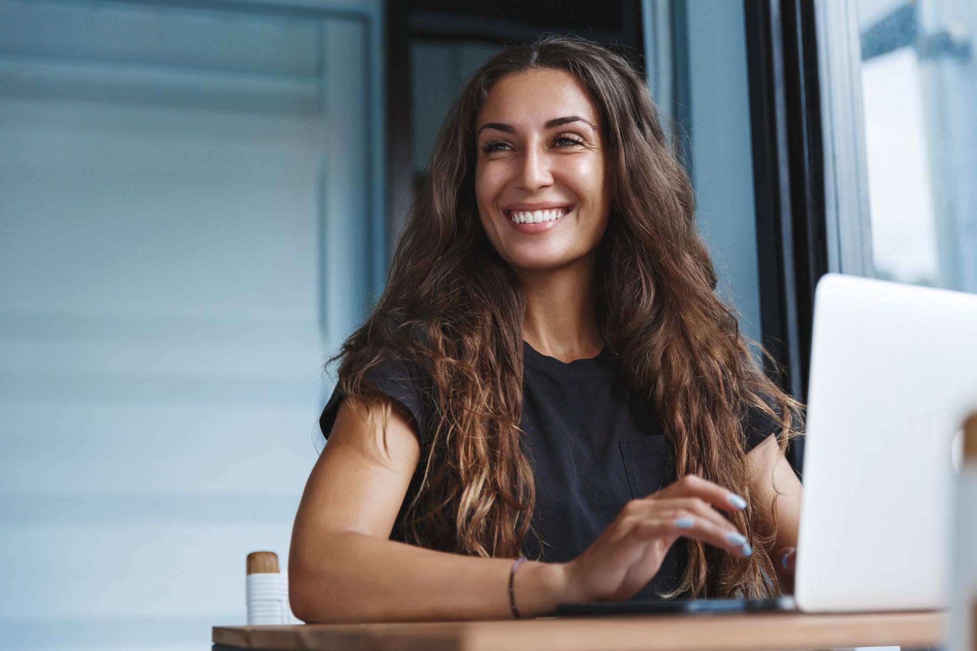 Woman with long, wavy brown hair smiles while working on a laptop at a table by a window.