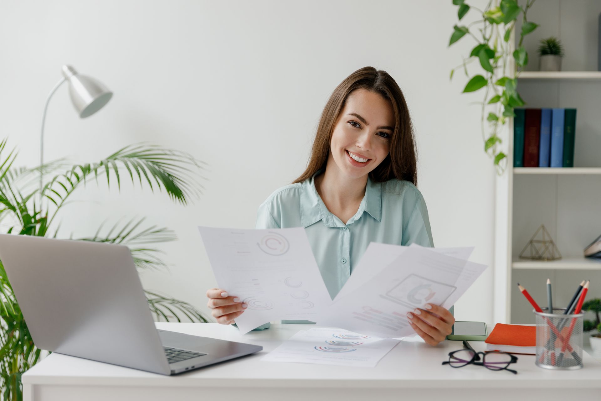 Woman smiles, holding papers, at desk with laptop, lamp, and plants. Woman smiles, holding papers, at desk with laptop, lamp, and plants.