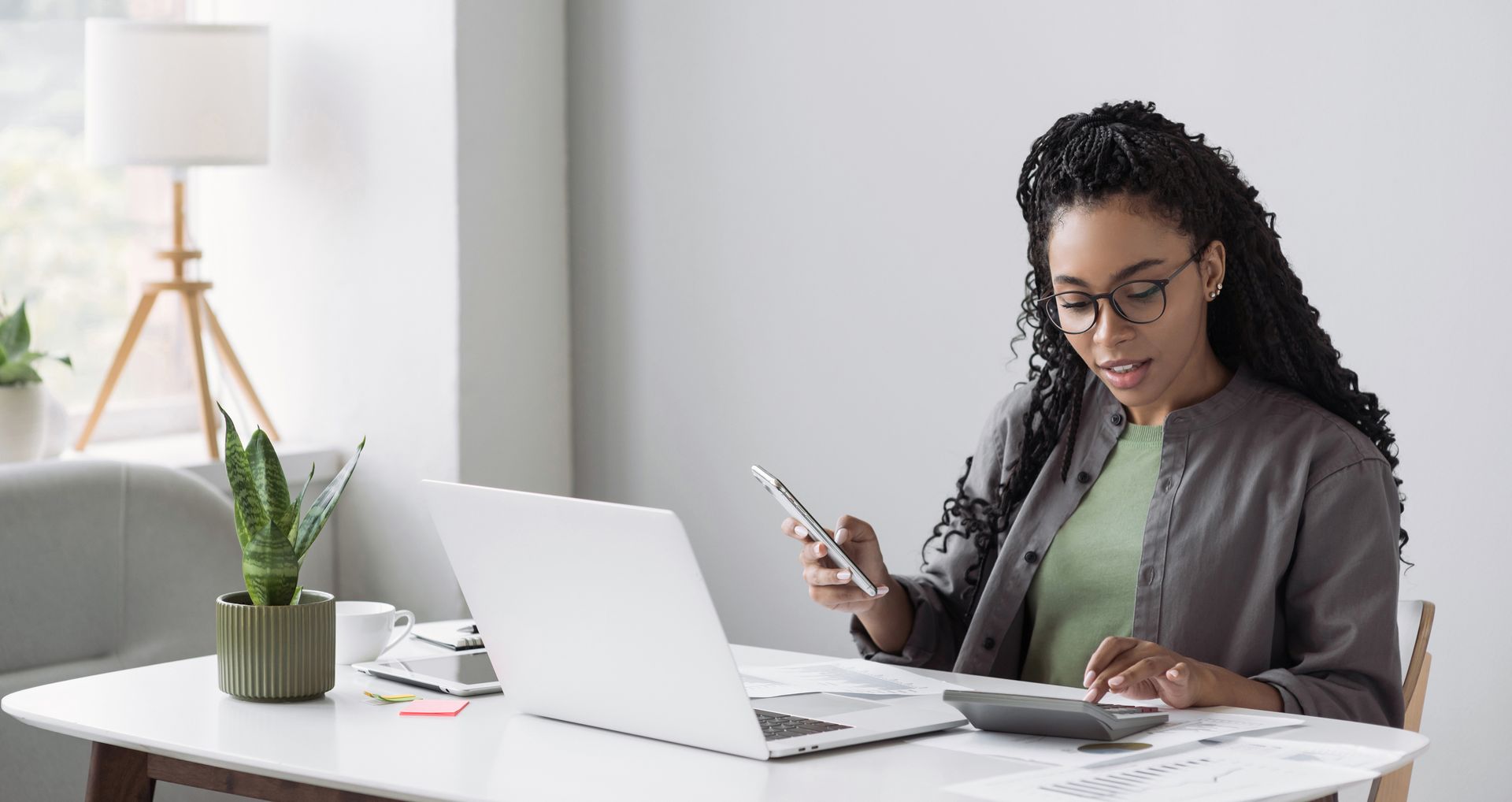 Woman wearing glasses using a calculator, laptop, and smartphone at a desk.