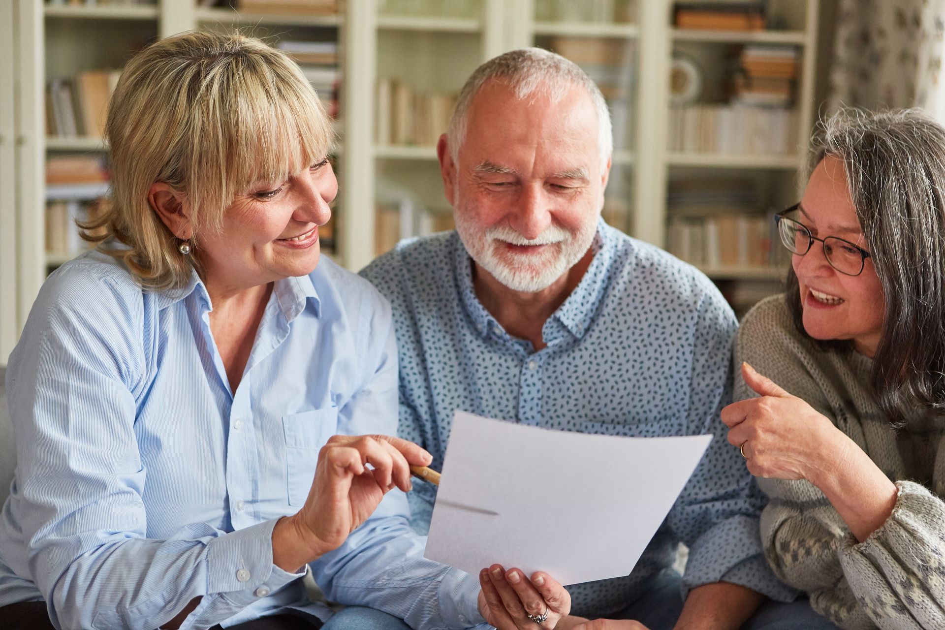 Three people looking at a document, smiling in a living room with bookshelves. Three people looking at a document, smiling in a living room with bookshelves.