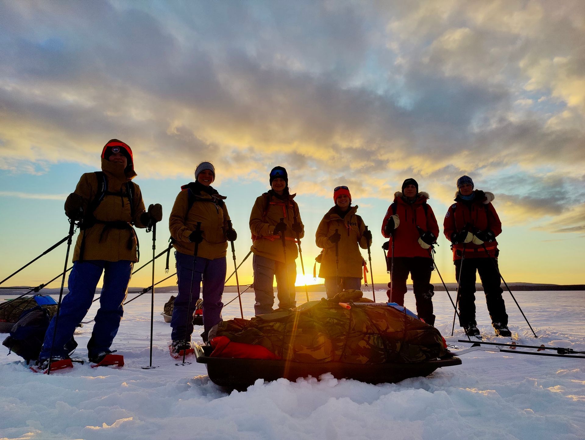 Six hikers with snowshoes stand on a snowy ridge at sunrise, backpacks and trekking poles in hand.