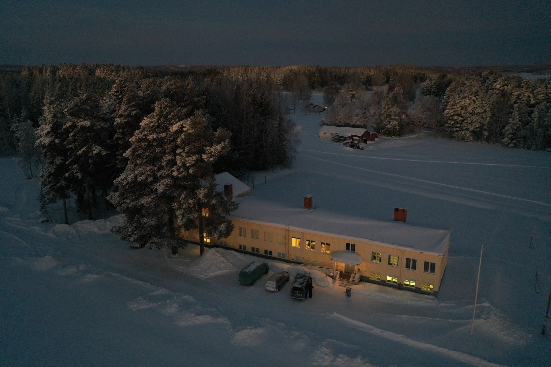 Snowy rural house lit warmly at dusk, with trees and fields covered in snow.